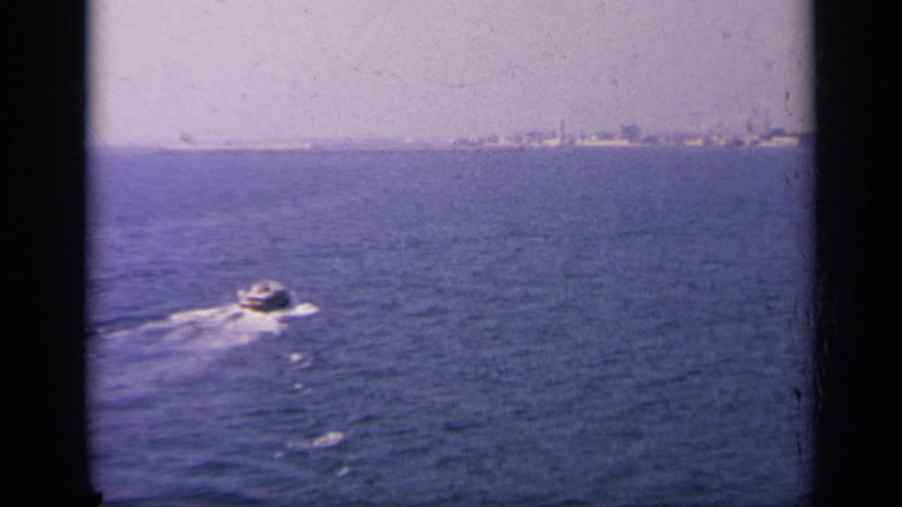 1963 Greece-area 8mm frame showing a motorboat leaving a wake across the sea, with a faint shoreline or harbor in the distance.