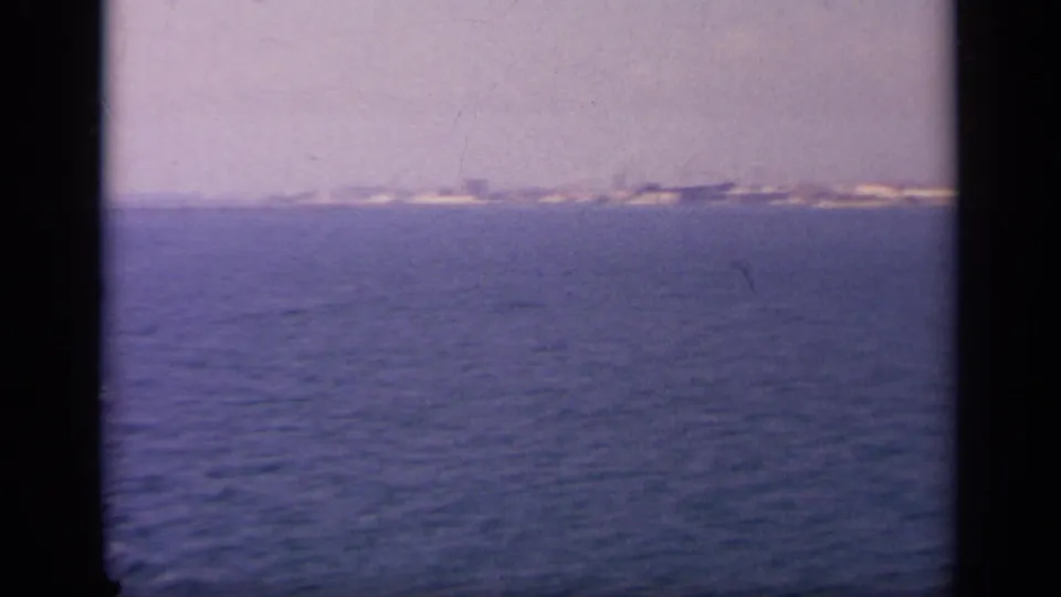 1963 Greece 8mm frame showing open blue water with a far white coastal town beneath a soft hazy sky.