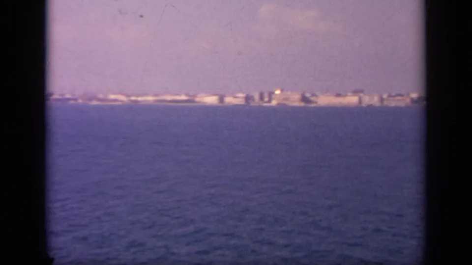 1963 Greece home-movie view of a low white coastal settlement seen from a boat across blue water.