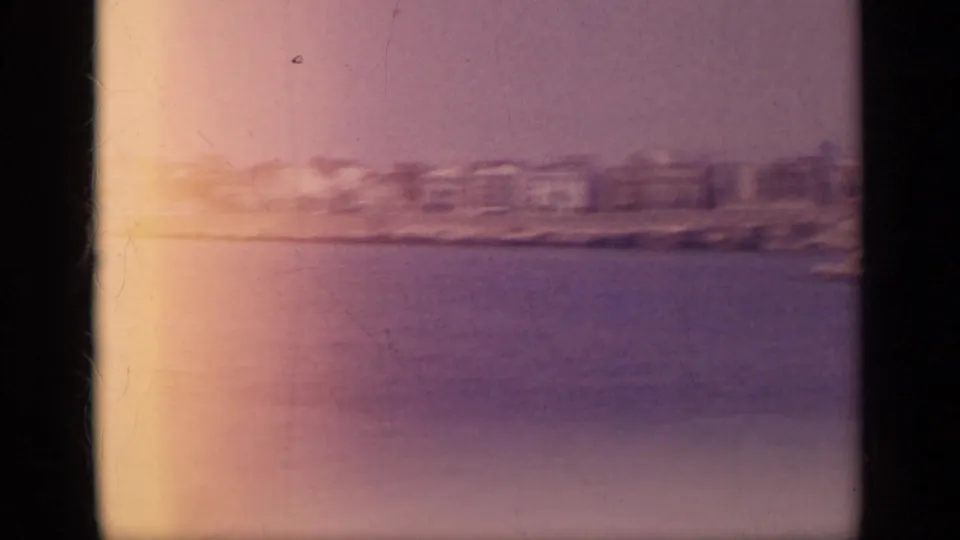 1963 Greece 8mm frame showing a softened shoreline across water, with warm exposure flare and visible film edge.