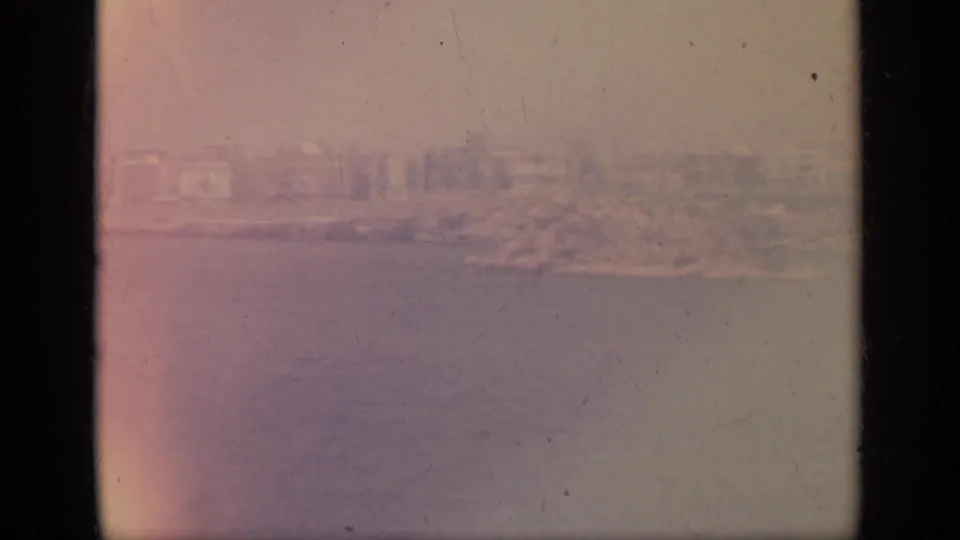 1963 8mm Greece footage showing a hazy coastal town or shoreline seen from the sea, with faded color and film wear.