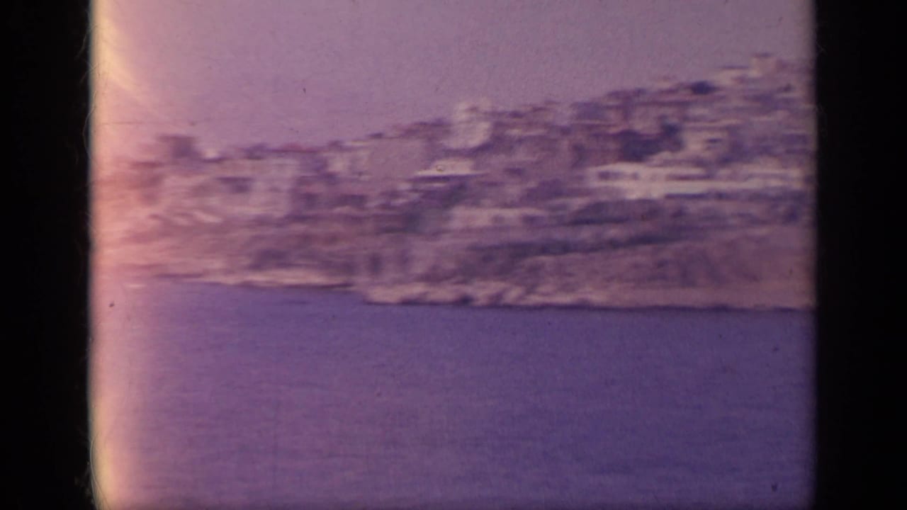 1963 8mm Greece view of a dense hillside coastal town overlooking blue sea, filmed from a moving boat.