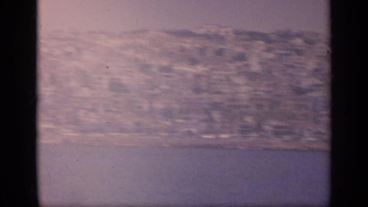 A soft, faded 1963 8mm Greece frame shows shoreline buildings and hillside terrain from offshore.
