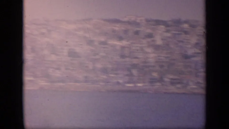 A soft, faded 1963 8mm Greece frame shows shoreline buildings and hillside terrain from offshore.