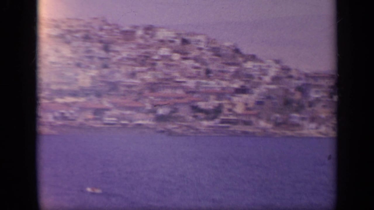 1963 8mm Greece travel footage frames a dense seaside hillside town, blue water, and aged magenta film color.
