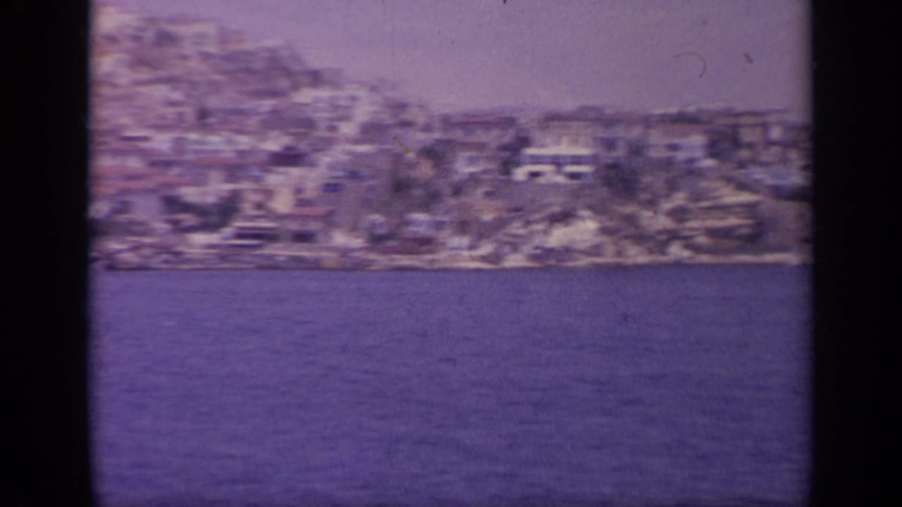 1963 Greek waterside town seen from across the water, with clustered hillside buildings and visible 8mm grain.