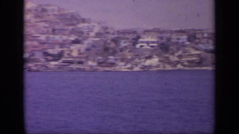 1963 Greek waterside town seen from across the water, with clustered hillside buildings and visible 8mm grain.