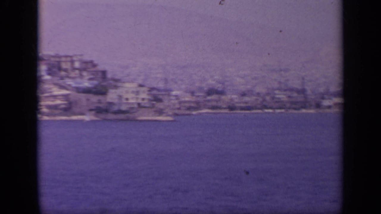 Coastal town continues along the water with dense pale buildings on a hillside.