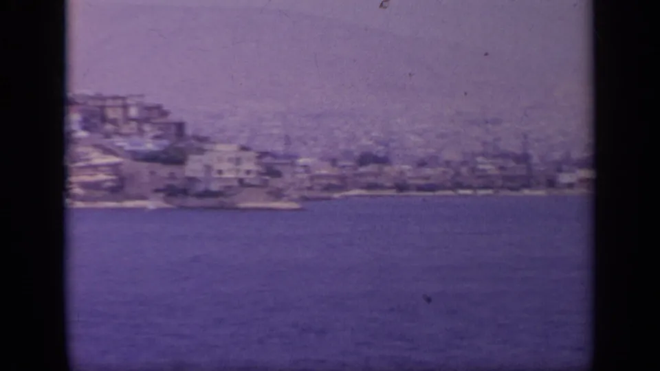 1963 Greece shoreline view with dense pale buildings along a hillside above the water, filmed from a distant vantage point.