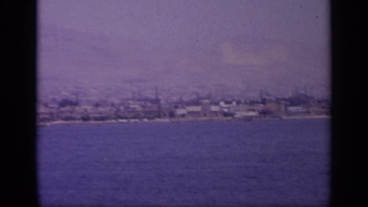 1963 Greek waterfront seen across open water, showing low shoreline buildings and possible industrial stacks in the distance.