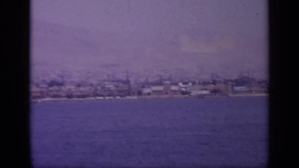 1963 Greek waterfront seen across open water, showing low shoreline buildings and possible industrial stacks in the distance.