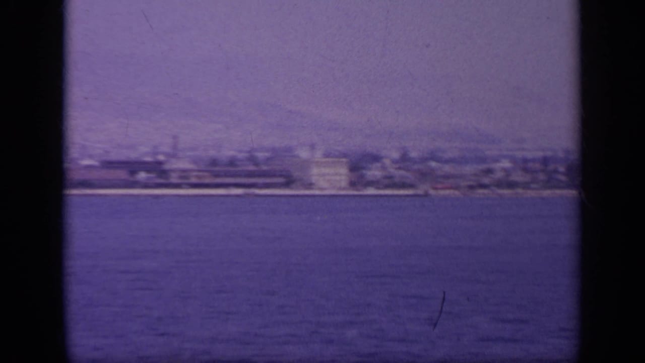 1963 Greece home-movie frame showing a hazy coastal city seen from a moving boat across open water.