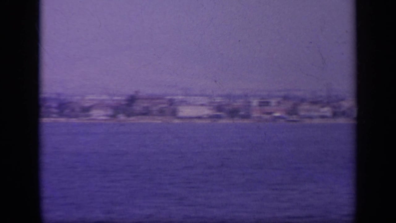 Soft 1963 Greek coastal view with low waterfront buildings and open water in the foreground.