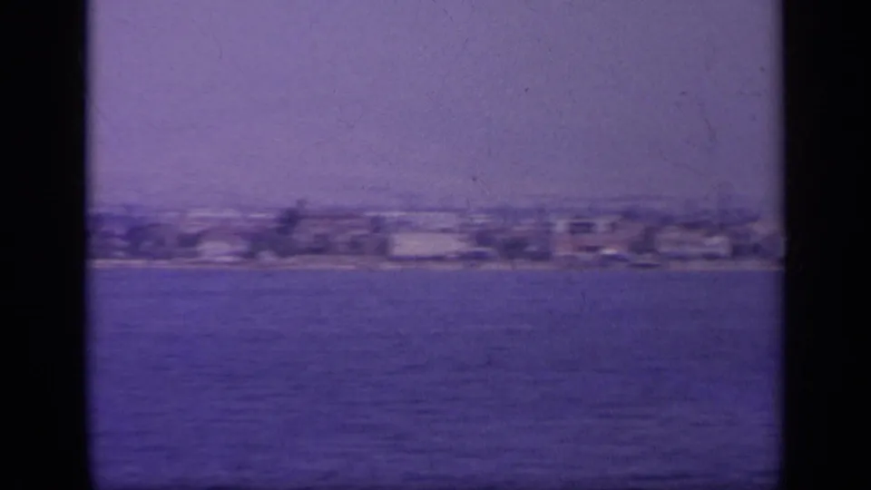 Soft 1963 Greek coastal view with low waterfront buildings and open water in the foreground.