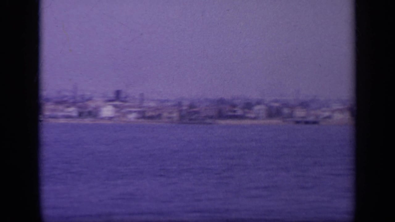 Aged 8mm frame of a Greek shoreline in 1963, with scattered waterfront structures seen from offshore.