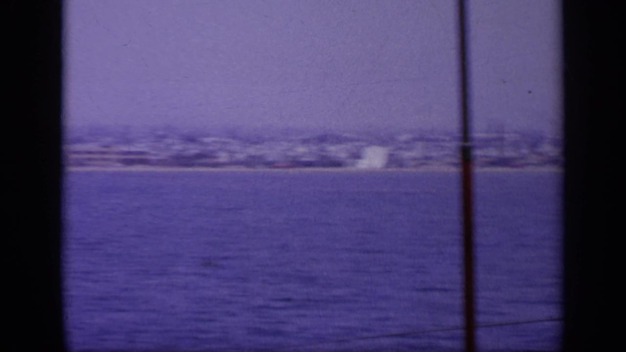 1963 Greece 8mm view across blue water toward a soft, hazy shoreline, partly obscured by dark vertical foreground bands.