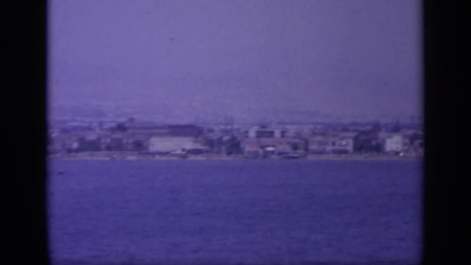 1963 Greece coastal view across blue water, with low waterfront buildings softened by haze and a dark band crossing the frame.