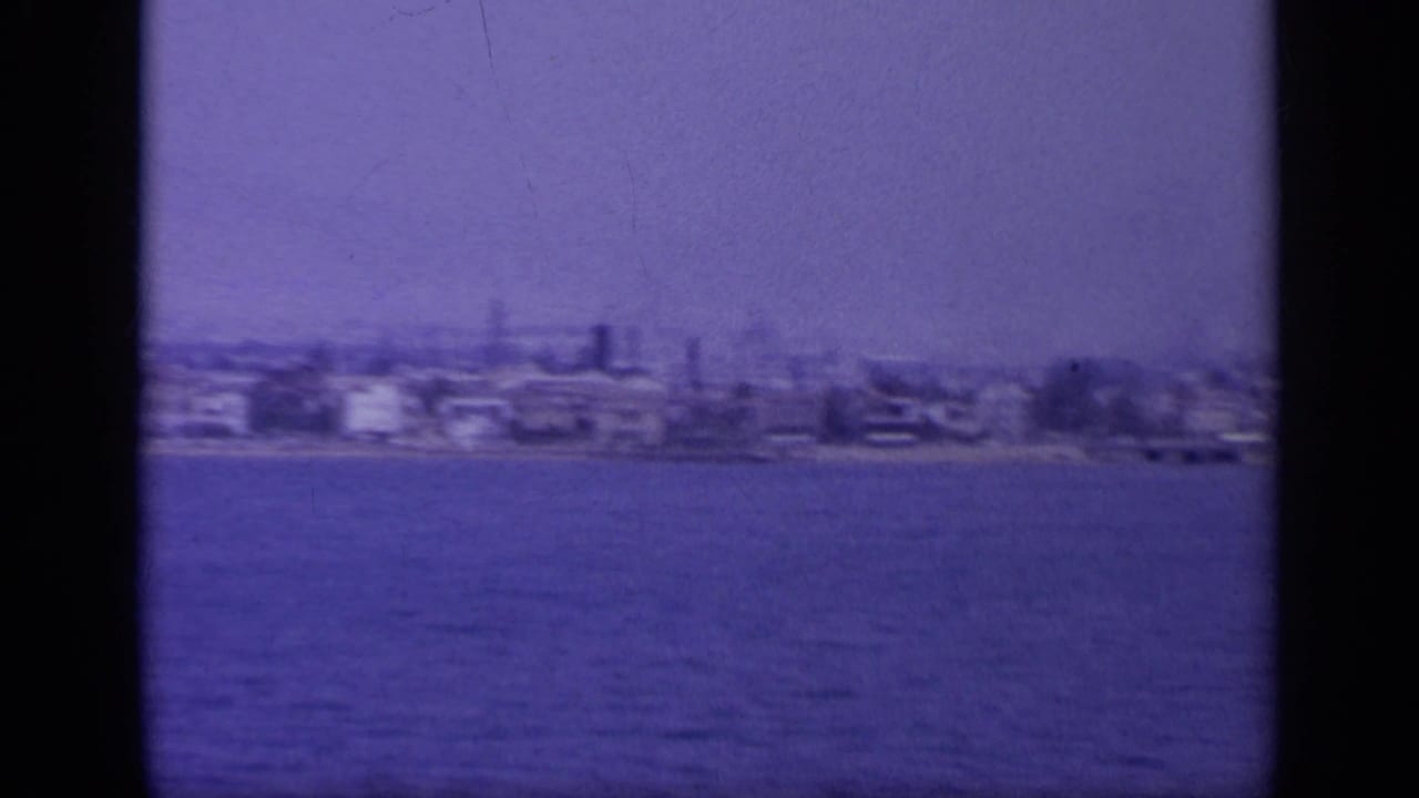 1963 Greece home-movie frame showing a distant coastal town across blue water, with soft focus, haze, and dark frame-edge bands.