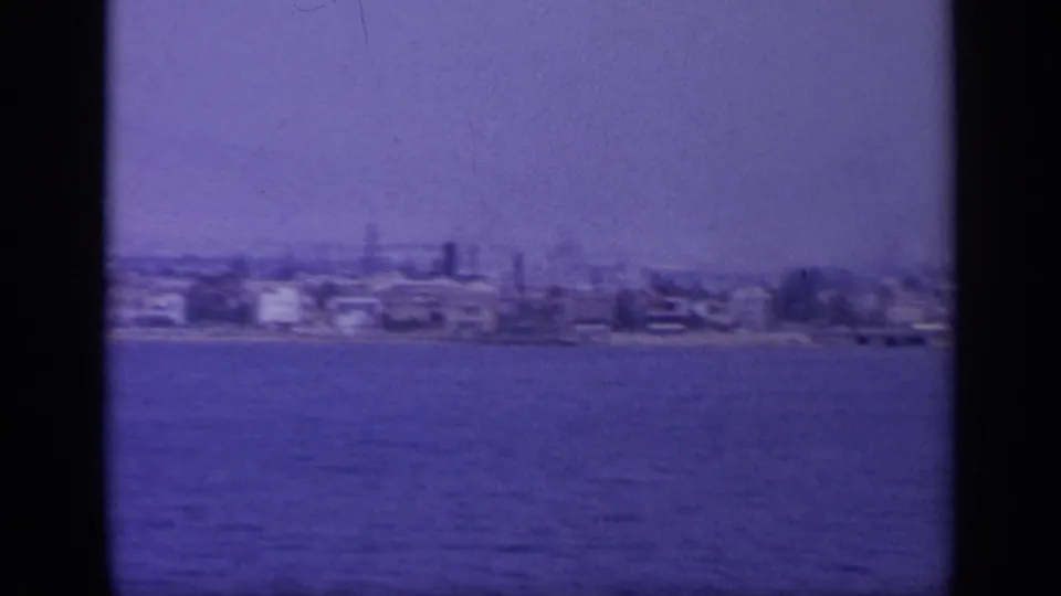1963 Greece home-movie frame showing a distant coastal town across blue water, with soft focus, haze, and dark frame-edge bands.