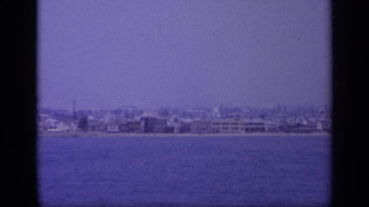 1963 Greece shoreline view across blue water, with scattered waterfront buildings, a pale hazy sky, and purple-blue 8mm color shift.