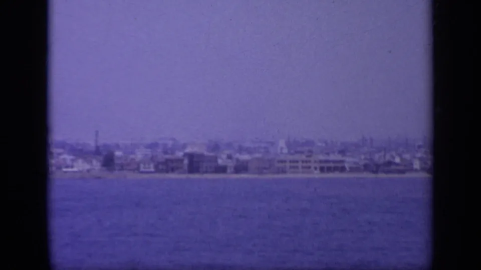 1963 Greece shoreline view across blue water, with scattered waterfront buildings, a pale hazy sky, and purple-blue 8mm color shift.