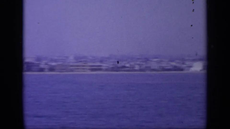 1963 8mm view of a Greek coastal city seen across water, with haze, soft focus, and dark frame borders.