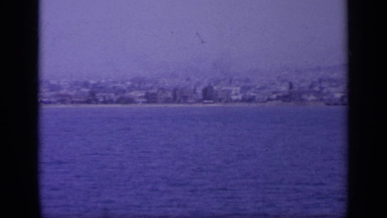 A soft 1963 home-movie frame shows buildings along a Greek shoreline beyond calm blue water.