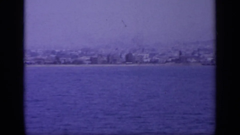 A soft 1963 home-movie frame shows buildings along a Greek shoreline beyond calm blue water.