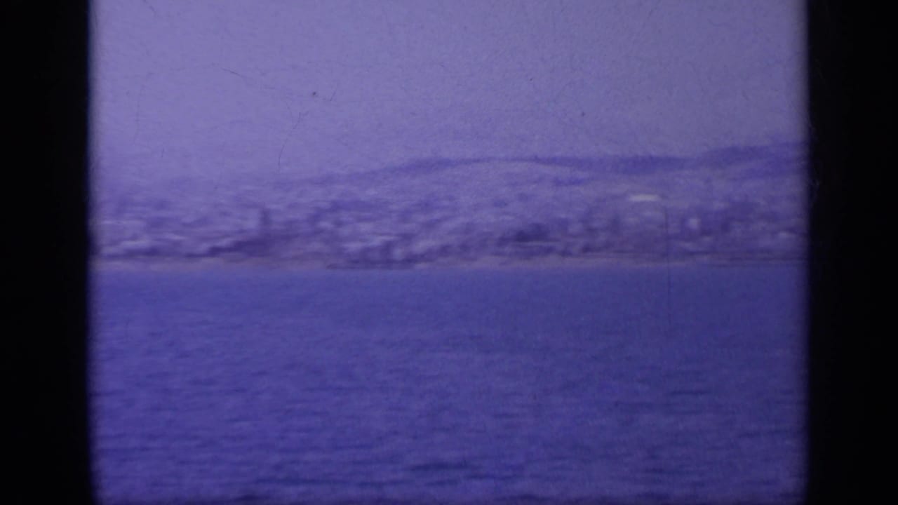 End of the pan across a Greek coastal view, with low hills, shoreline buildings, and blue water in 8mm texture.