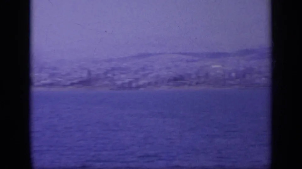 End of the pan across a Greek coastal view, with low hills, shoreline buildings, and blue water in 8mm texture.