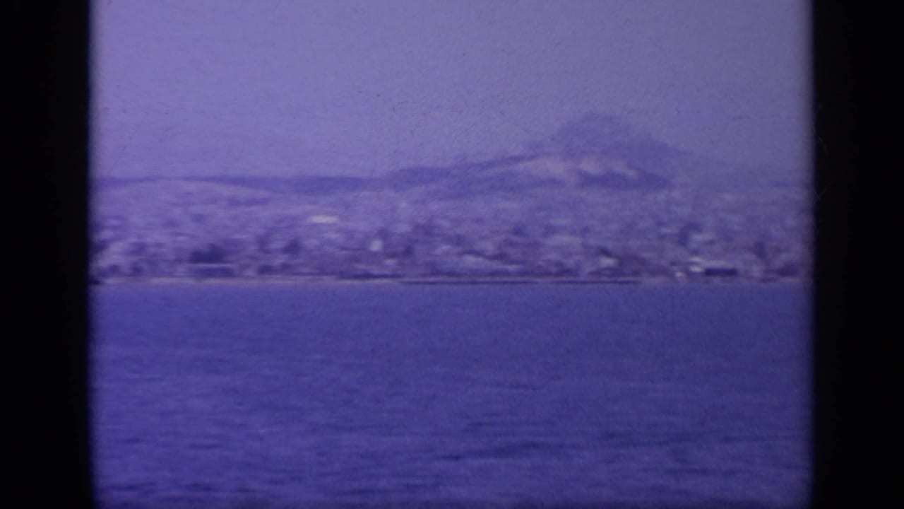 1963 8mm view across water toward a Greek coastal town and hazy mountains, tinted blue by aged film stock.