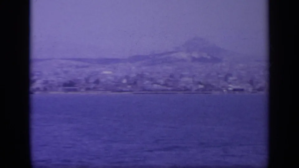1963 8mm view across water toward a Greek coastal town and hazy mountains, tinted blue by aged film stock.