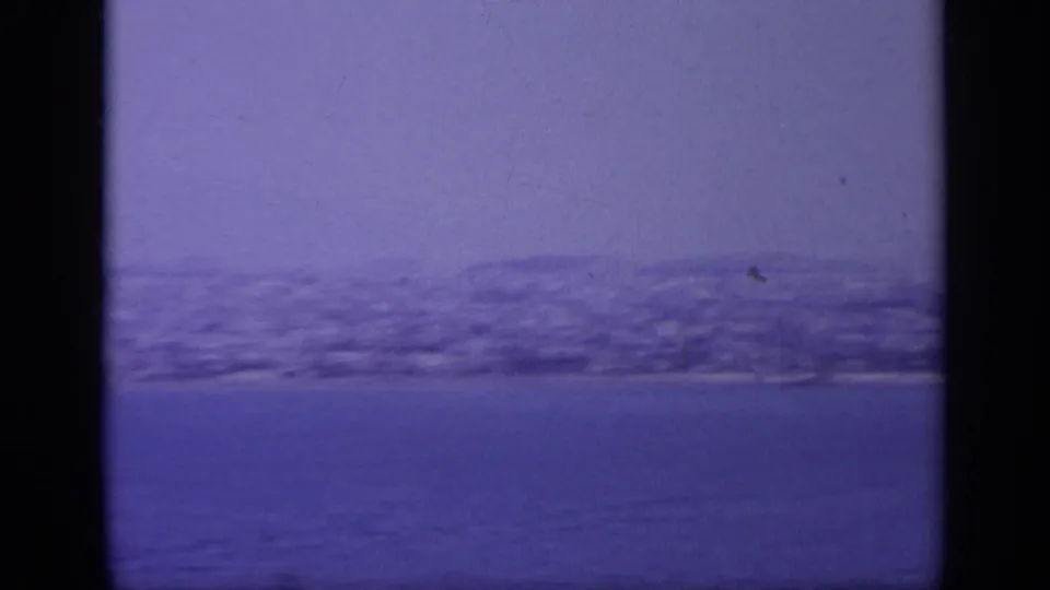 Blue-tinted 1963 8mm frame showing a distant Greek shore, low hills, and soft horizon haze beyond the water.