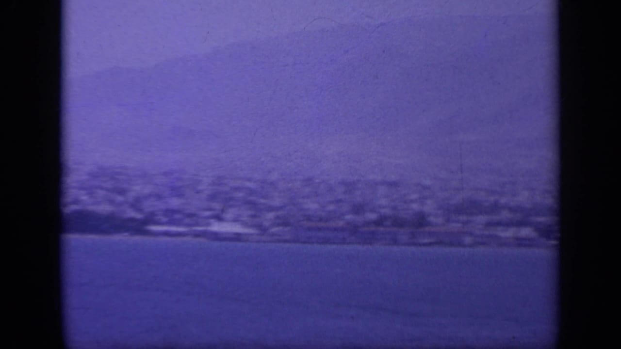 1963 home-movie frame of a Greek coastal settlement below hills, seen across calm water with visible 8mm scan borders.
