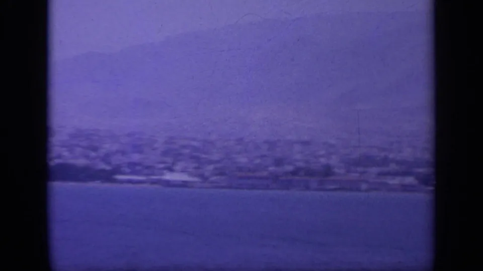 1963 home-movie frame of a Greek coastal settlement below hills, seen across calm water with visible 8mm scan borders.