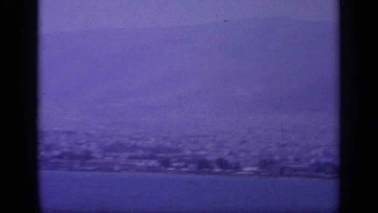 1963 8mm view of a Greek shoreline seen from offshore, with hazy hills and water under a purple-blue film cast.