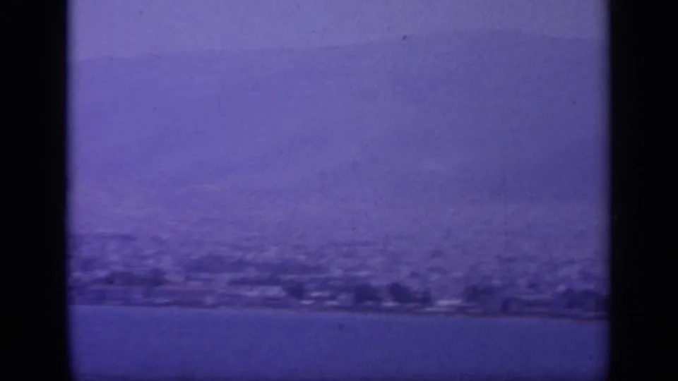 1963 8mm view of a Greek shoreline seen from offshore, with hazy hills and water under a purple-blue film cast.