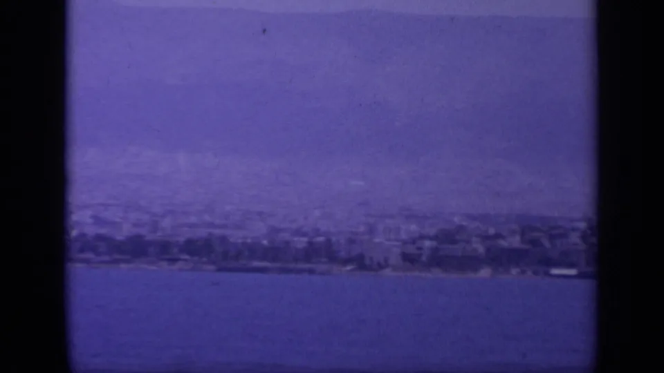 Greek coastal waterfront seen from a moving boat, with distant buildings, open sea, and hazy mountains in 1963 8mm footage.