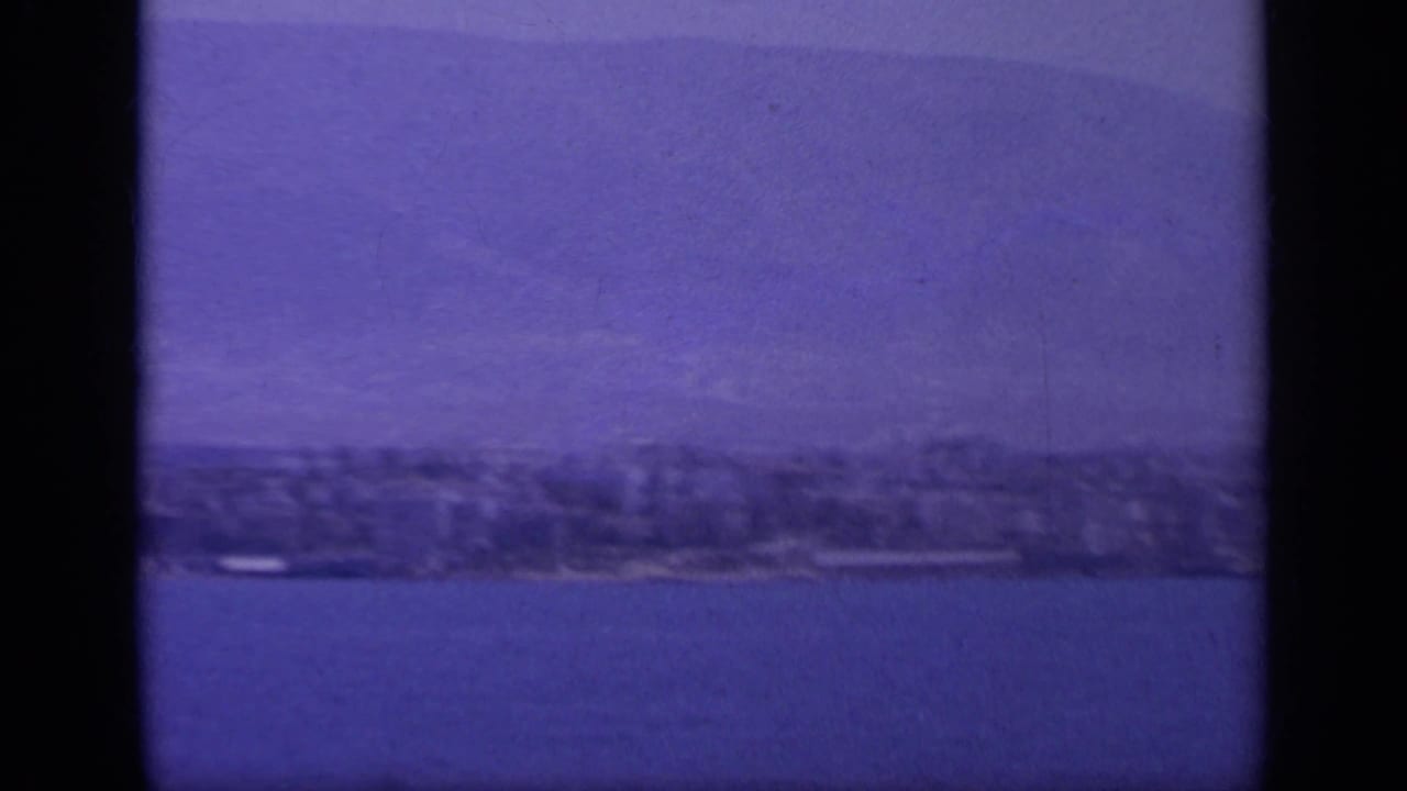 Offshore 8mm travel view of a Greek coast, showing blue water, soft-focus shoreline, and pale mountains in the distance.
