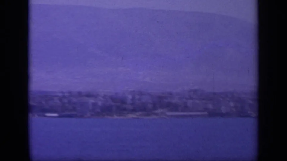 Offshore 8mm travel view of a Greek coast, showing blue water, soft-focus shoreline, and pale mountains in the distance.