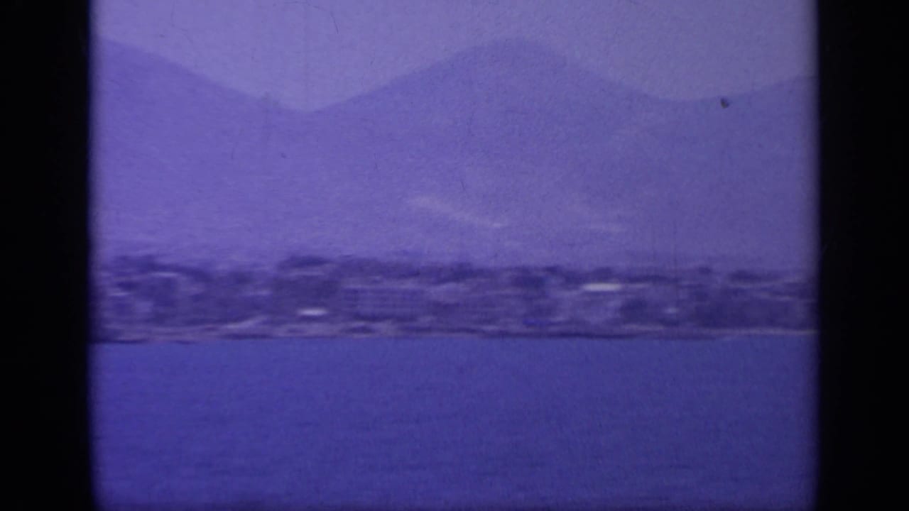 1963 Greek coastal cruise frame with water in the foreground, a low shoreline, and layered mountains behind it.