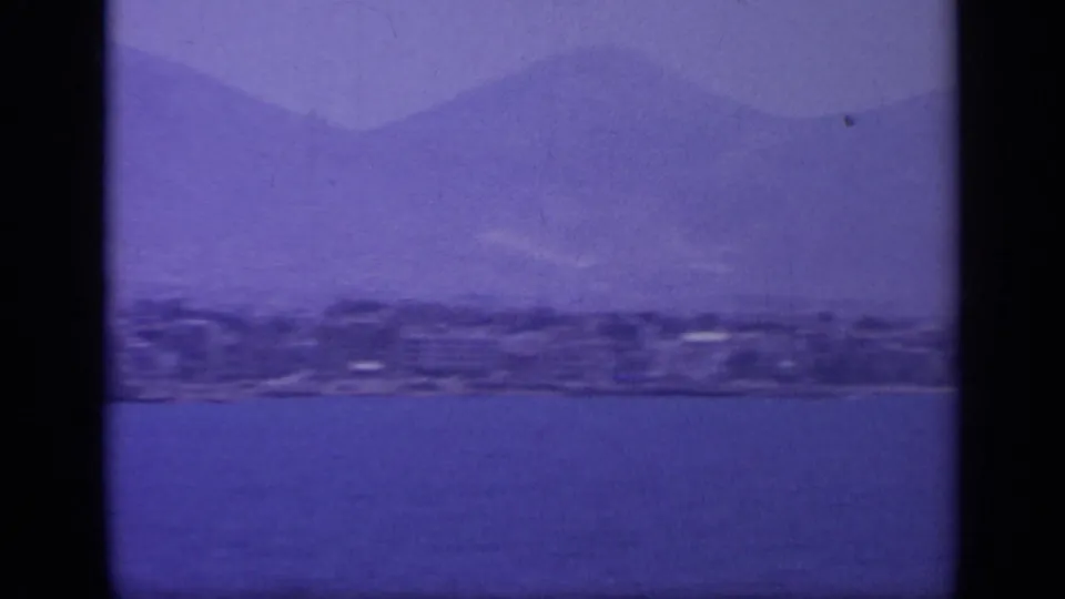 1963 Greek coastal cruise frame with water in the foreground, a low shoreline, and layered mountains behind it.