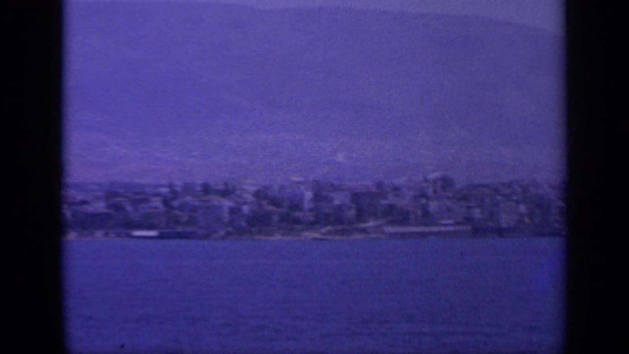 1963 Greece 8mm view of a shoreline town across calm sea, with hazy mountain ridges and dark film-frame edges.