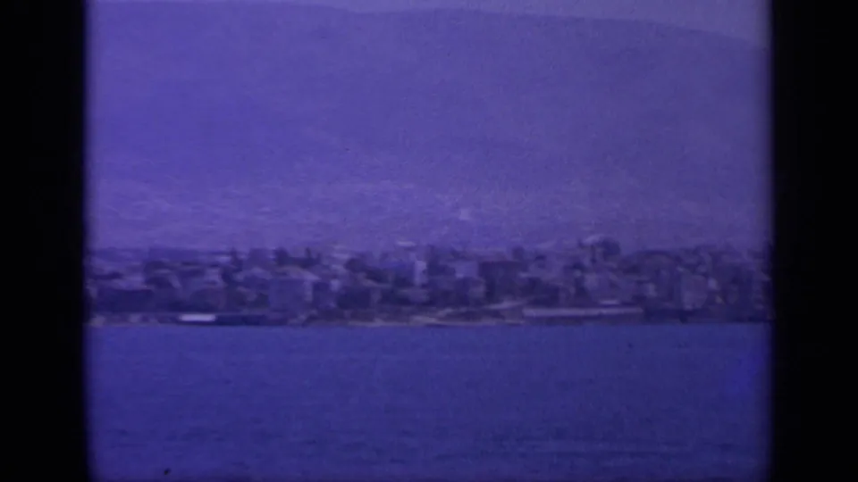 1963 Greece 8mm view of a shoreline town across calm sea, with hazy mountain ridges and dark film-frame edges.
