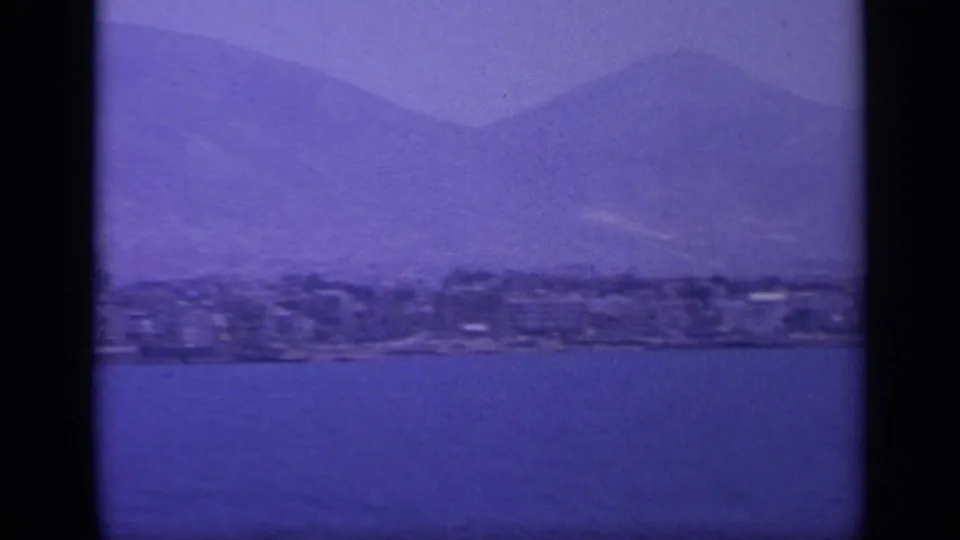 1963 Greece coastal panorama with buildings along the shore, sea in the foreground, and soft mountains beyond.