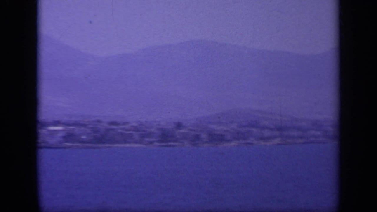 Aged 8mm frame from 1963 Greece showing low shoreline land, calm water, and layered mountains in blue-purple tones.