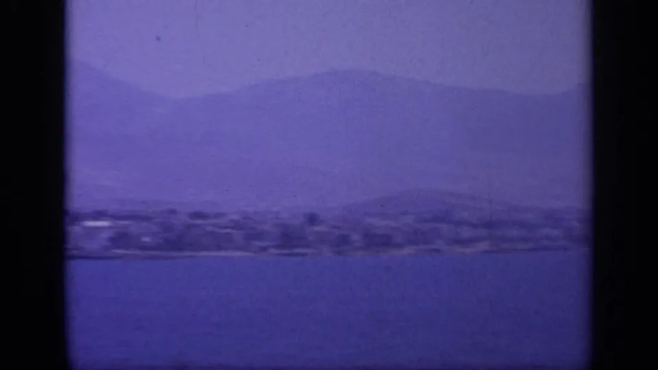 Aged 8mm frame from 1963 Greece showing low shoreline land, calm water, and layered mountains in blue-purple tones.