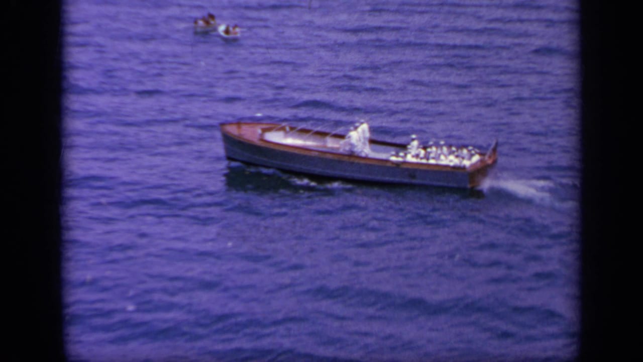 1963 Greece home-movie frame of a wooden launch with uniformed men and a small boat farther across the water.
