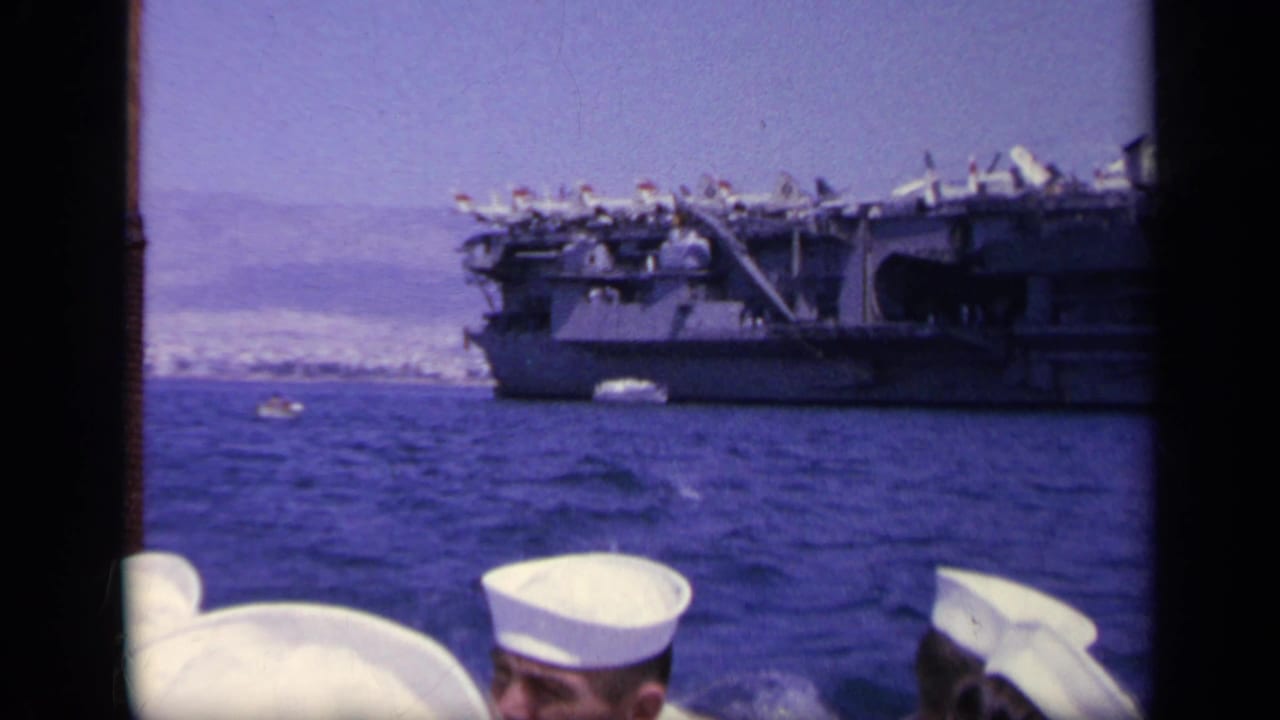 1963 Greece 8mm frame showing a larger naval vessel with aircraft on deck, viewed past sailors' white hats from another boat.