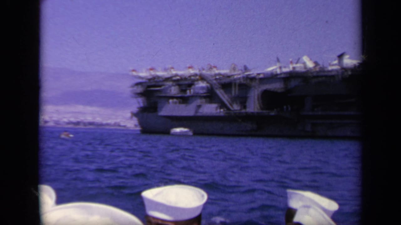 Sailors' white caps in foreground with an aircraft carrier across blue water.